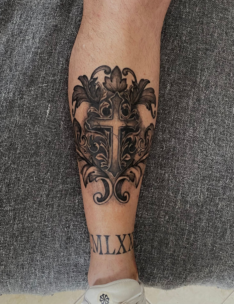 Decorative black tattoo on a person's leg of Roman numerals and cross, Almancil.