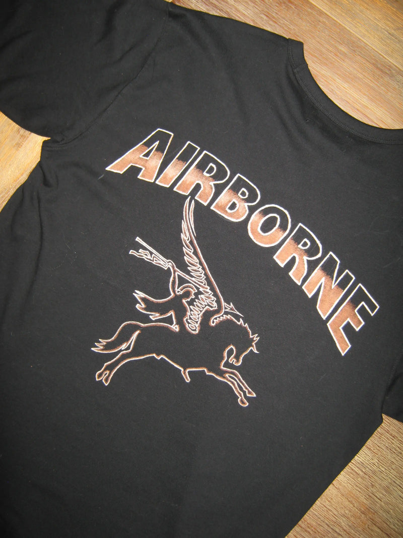 Black t-shirt with 'AIRBORNE' text and a pegasus on a wooden surface